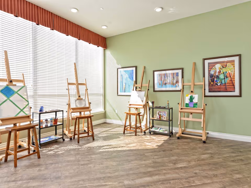 A bright art studio room with large windows covered by blinds and a red valance. The room has light green walls and wooden flooring. There are five wooden easels, each holding a canvas with various paintings or sketches. Two black rolling carts hold art supplies such as paint bottles and brushes. Three framed artworks hang on the wall.