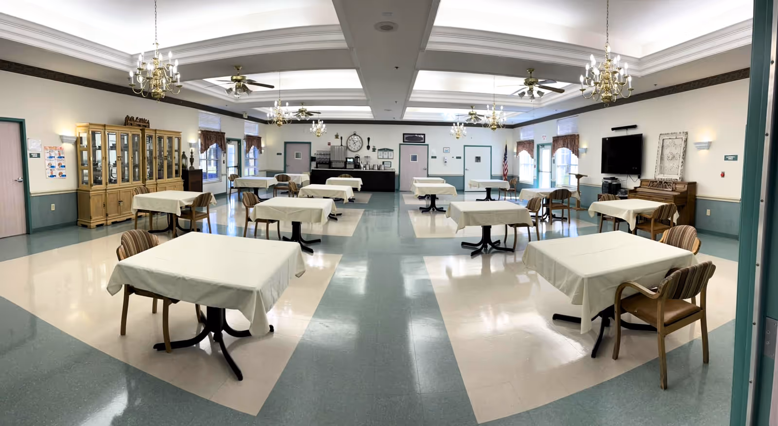A spacious dining room with multiple tables covered in white tablecloths, each surrounded by wooden chairs with striped cushions. The room features a high ceiling with chandeliers and ceiling fans, large windows with valances, a wooden cabinet on the left, and a wall-mounted TV on the right. The floor has a patterned tile design and the walls are painted in light colors with wainscoting.