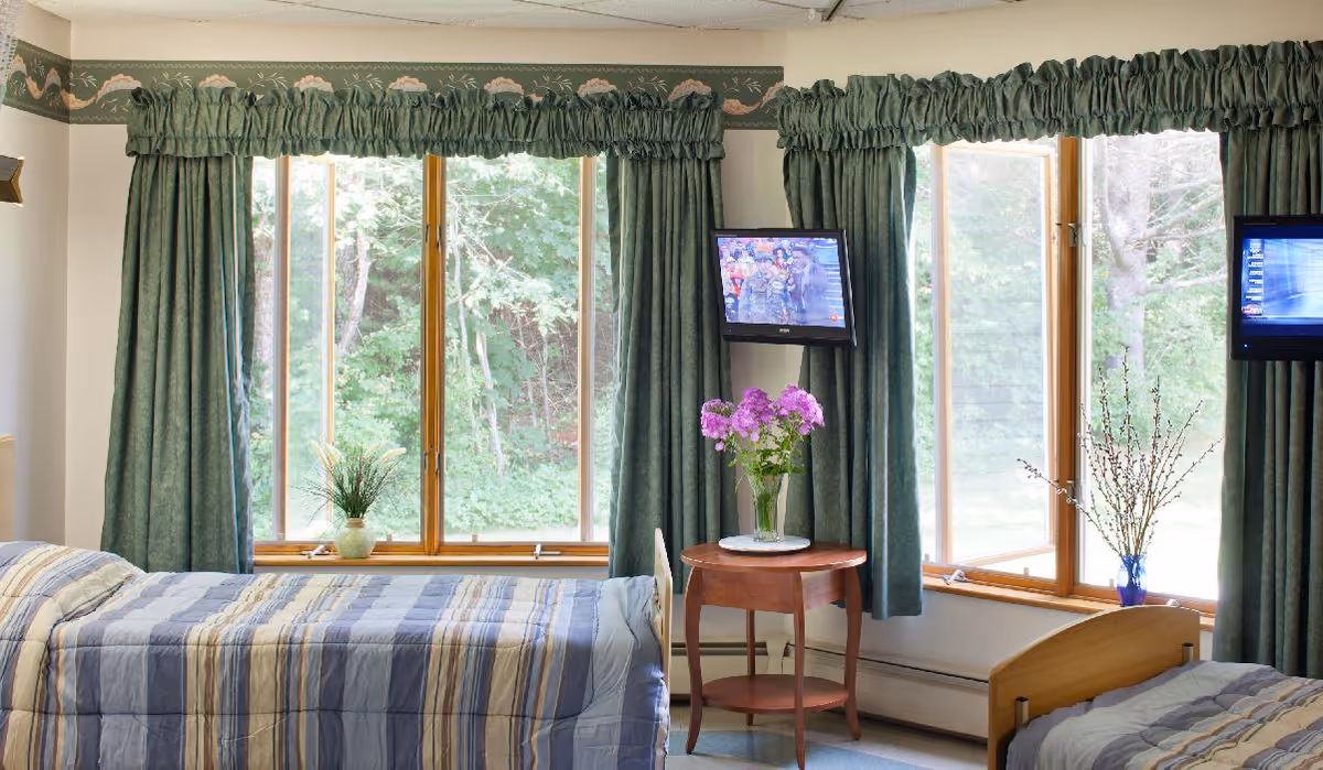 A bright room with two single beds covered in striped blue and beige bedding. Between the beds is a small round wooden table with a vase of purple flowers. The room has large windows with green curtains and valances, showing a view of green trees outside. Two small wall-mounted televisions are visible above each bed.
