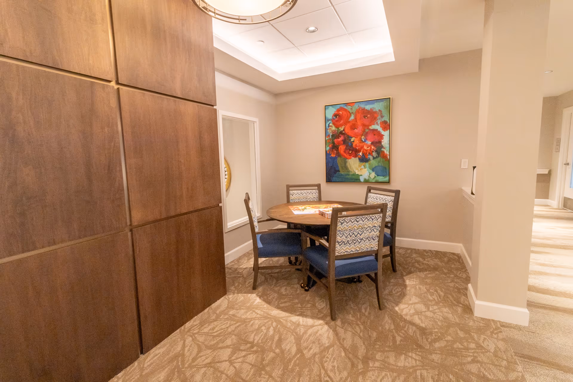 Small seating area with a round wooden table and four upholstered chairs beneath ceiling lighting and a colorful floral painting on the wall.