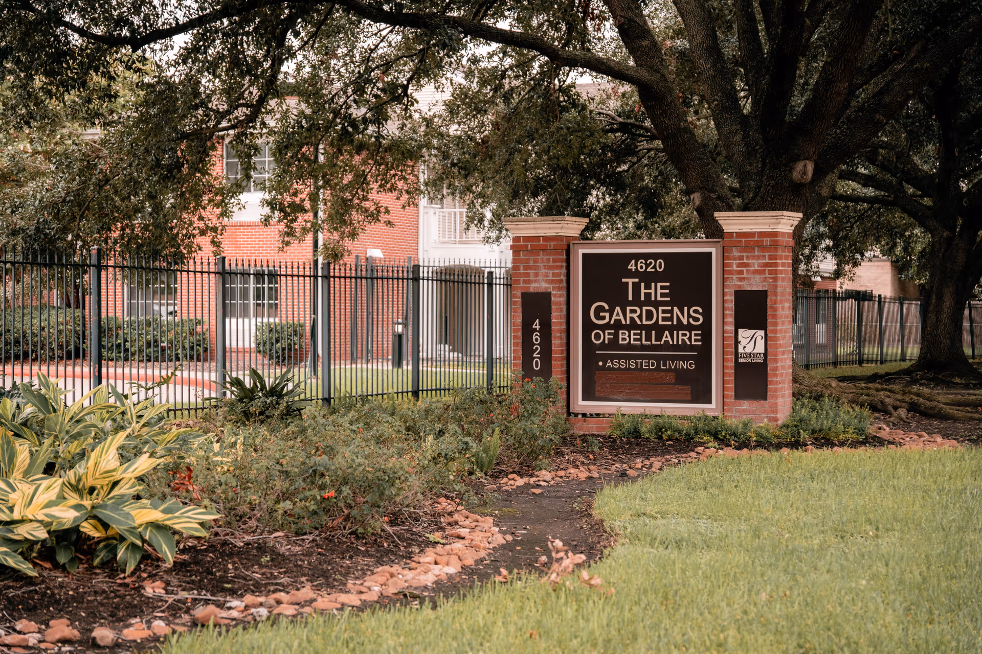 Outdoor view of the entrance sign for The Gardens of Bellaire assisted living facility, featuring a brick sign with the address 4620, surrounded by greenery, plants, and large trees with a black metal fence and a red brick building in the background.