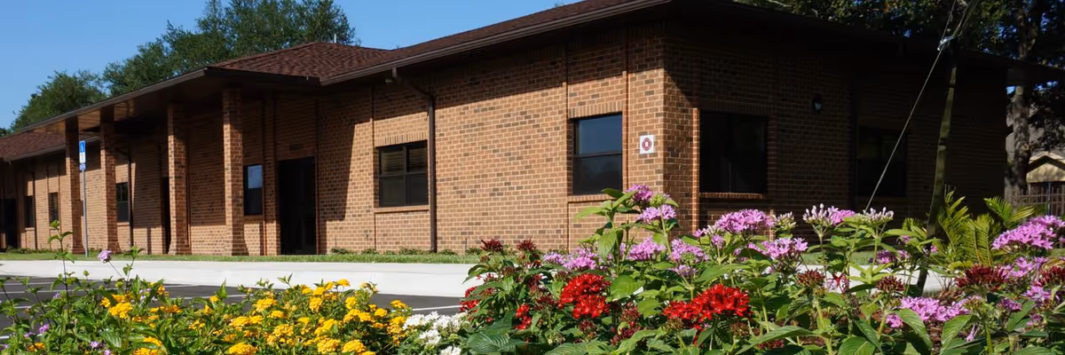 Single-story brick building with windows and an entrance behind a colorful flower bed under a clear blue sky.
