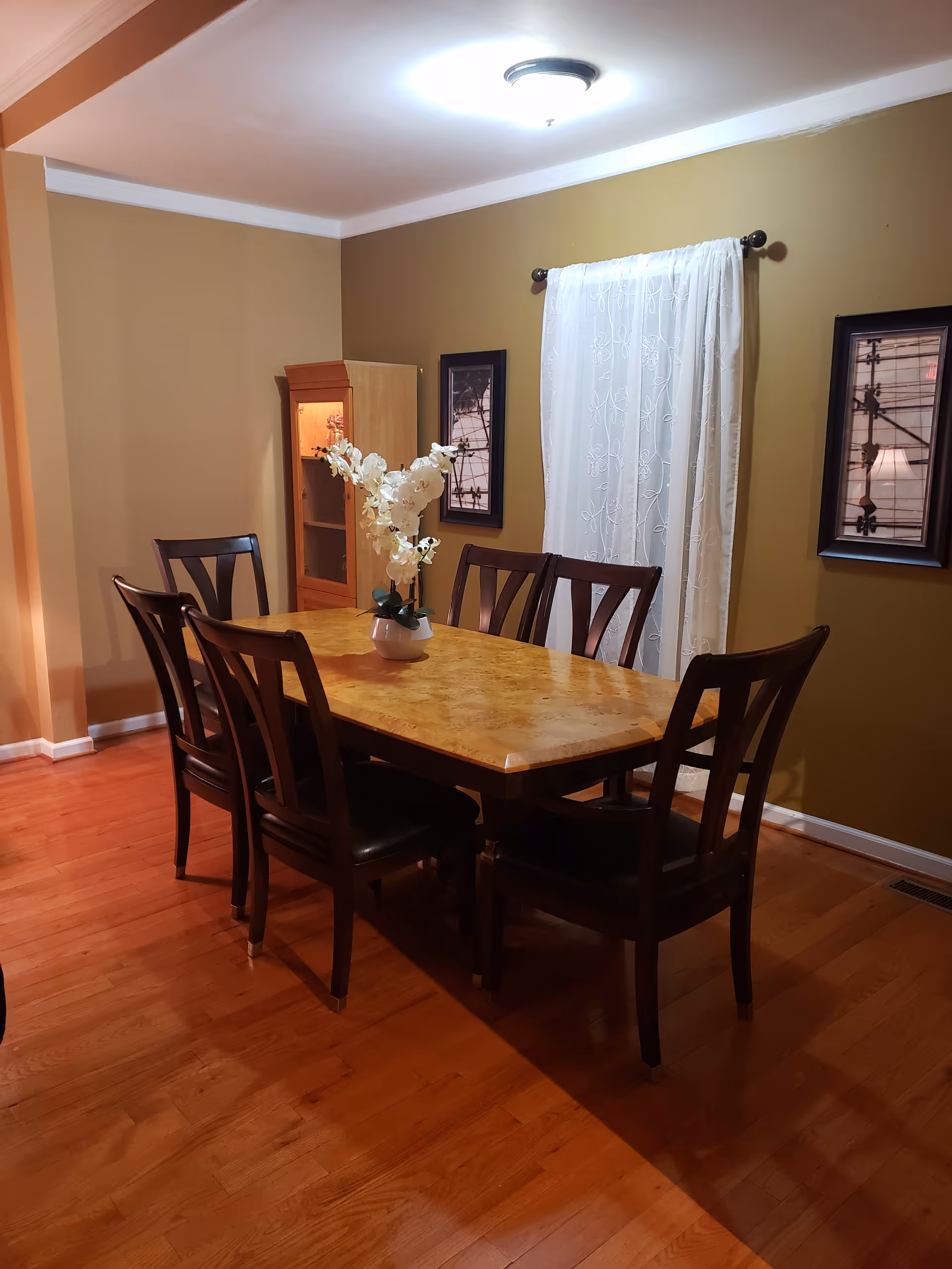 A dining room with a wooden dining table surrounded by six dark wooden chairs with black cushions. A white vase with white flowers is placed in the center of the table. The room has wooden flooring, beige walls, and a window with white sheer curtains. Two framed pictures hang on the wall on either side of the window. A wooden cabinet is positioned in the corner of the room.