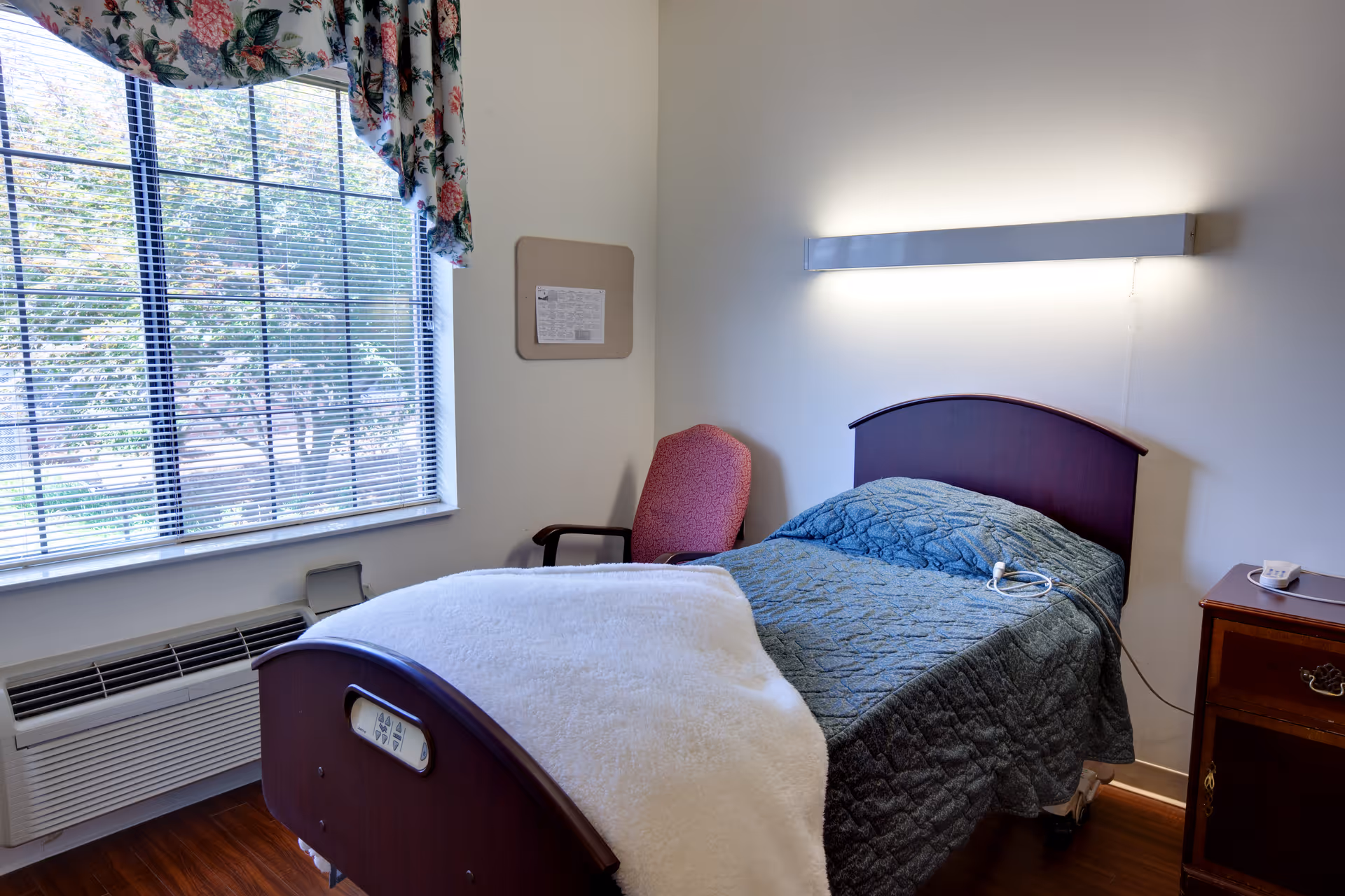 A small bedroom in a senior living facility with a single bed covered with a blue quilt and a white blanket. There is a red upholstered chair next to the bed, a wooden nightstand with a white cup, and a large window with floral curtains letting in natural light. A long horizontal light fixture is mounted on the wall above the bed.