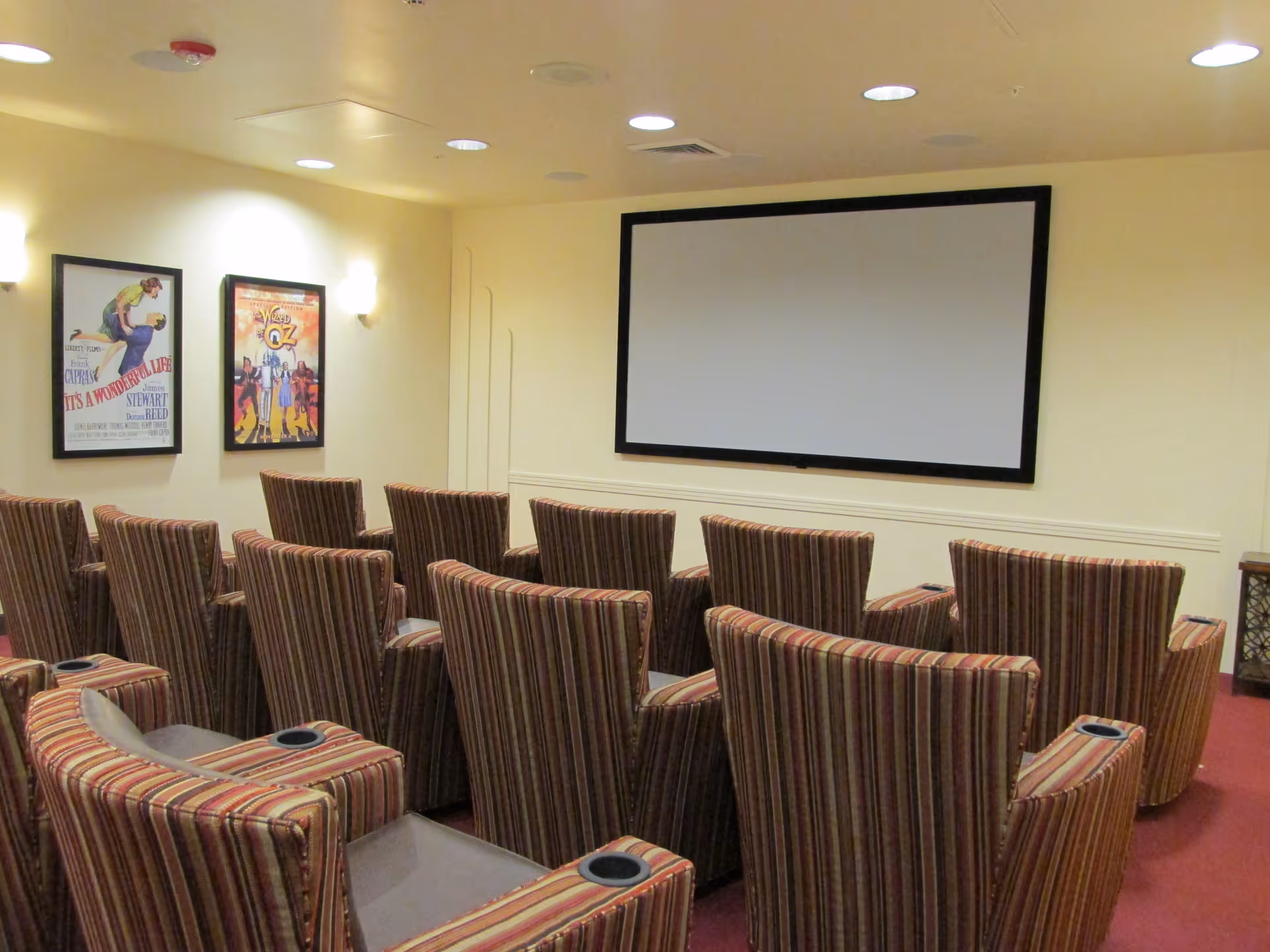 A small theater room with multiple striped upholstered chairs arranged in rows facing a large blank projection screen on the wall. Two framed movie posters are hung on the left wall, and the room is softly lit with wall sconces.