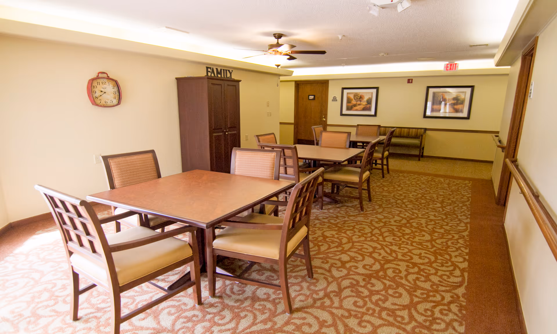 A well-lit room with multiple square wooden tables and cushioned chairs arranged for seating. The room has patterned carpet flooring, a ceiling fan with lights, framed artwork on the walls, a wooden cabinet with the word FAMILY on top, and an exit sign above a door in the background.