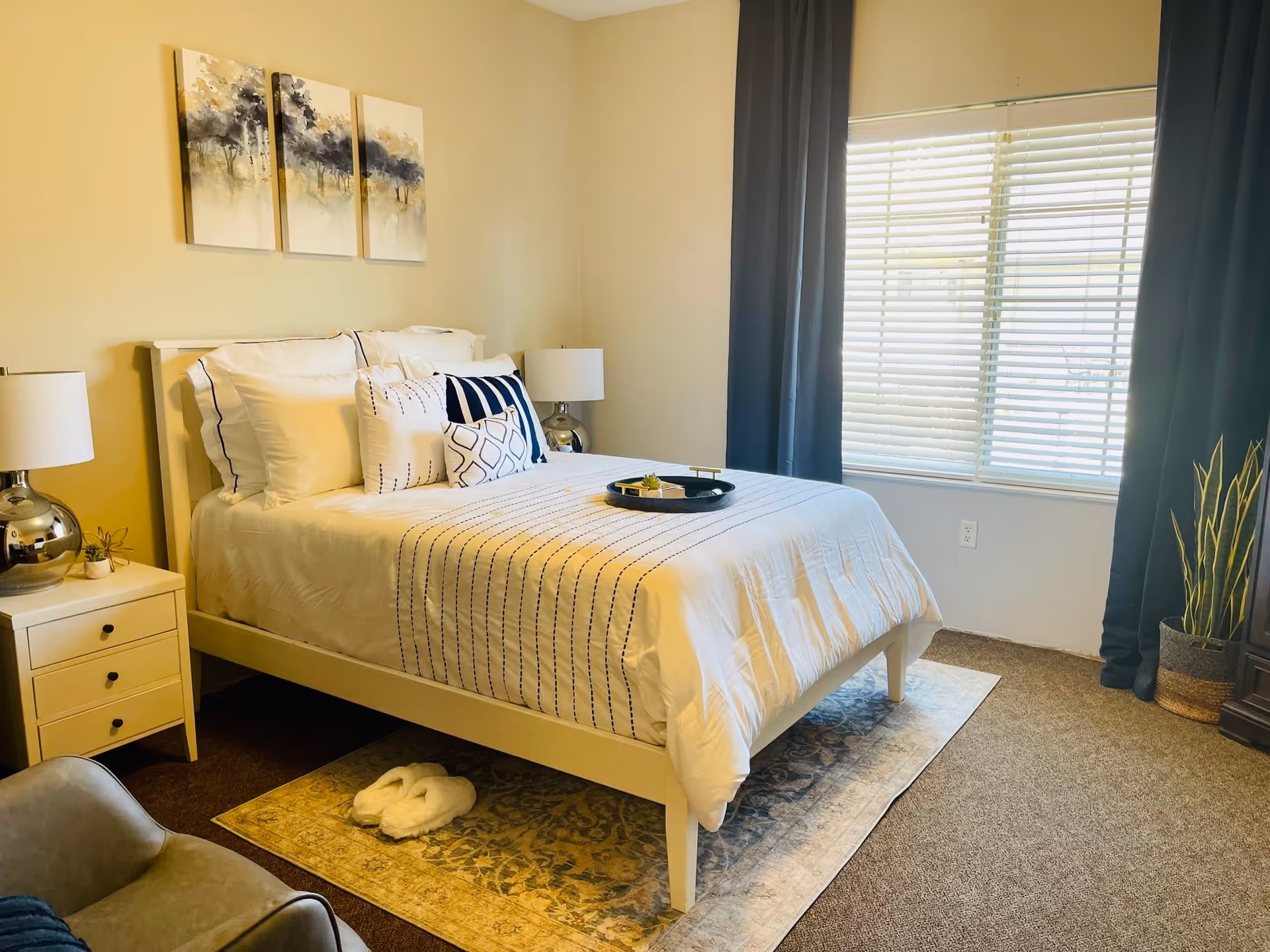 A neatly made bed with white and blue striped bedding and multiple pillows in a bedroom. There are two bedside tables with lamps on each side of the bed. A pair of white slippers is placed on a patterned rug under the bed. The room has a large window with white blinds and dark curtains, a potted plant in the corner, and three abstract paintings on the wall above the bed.
