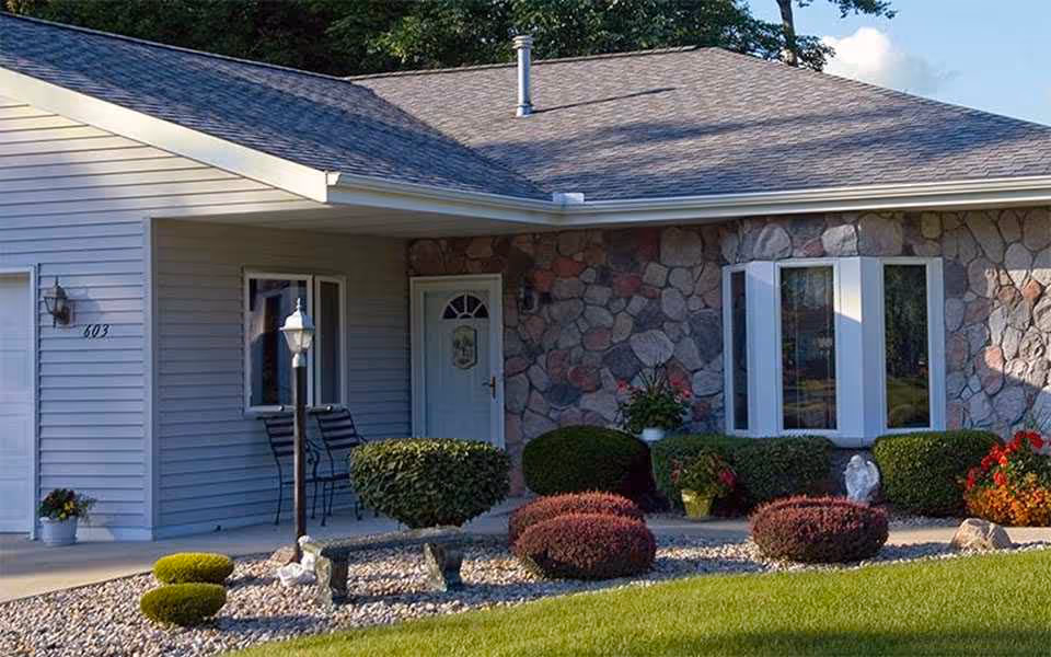 Exterior view of a single-story retirement community unit with a stone and siding facade, a white front door, a bay window, and a small porch area with two chairs. The front yard is landscaped with trimmed bushes, a stone bench, a lamp post, and colorful flowers.