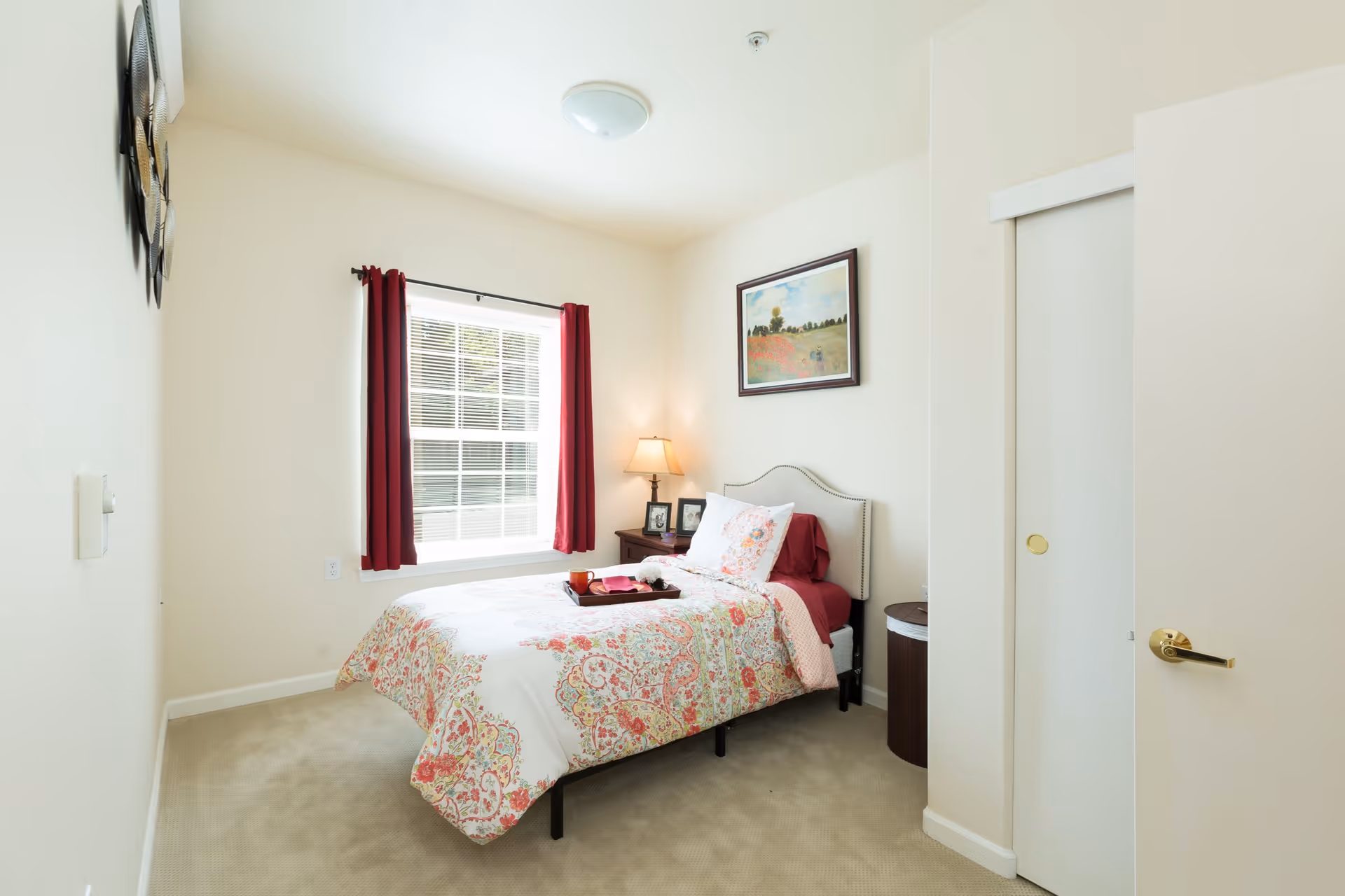Bright single bedroom with a floral-covered bed, window with red curtains, nightstand and lamp, and framed artwork on the wall.
