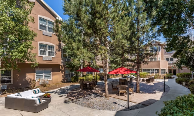Sunny courtyard with outdoor seating, red umbrellas, trees and surrounding apartment building.