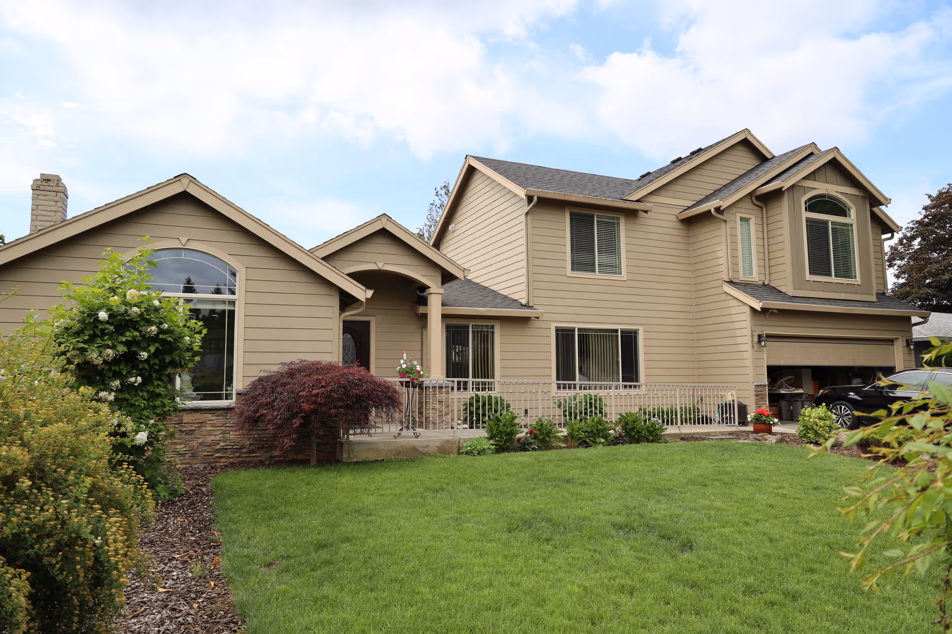 Exterior view of a two-story beige house with a well-maintained green lawn and various shrubs and plants in the front yard under a partly cloudy sky.