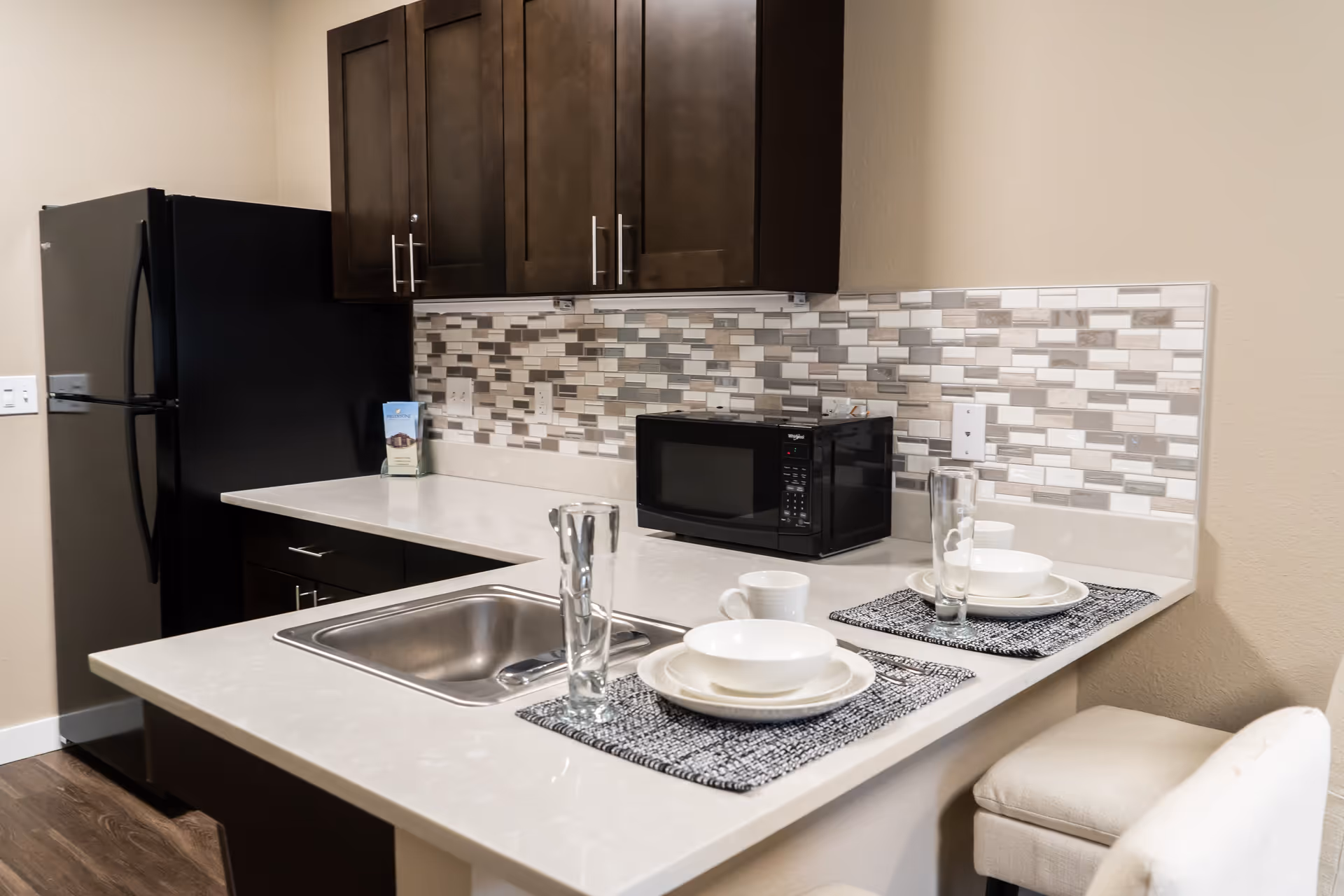 A modern kitchen area with dark wooden cabinets, a black refrigerator, a black microwave on the countertop, and a stainless steel sink. The backsplash features a mosaic tile pattern in shades of gray, white, and beige. Two place settings with white dishes, cups, and tall glasses are arranged on the counter with gray placemats. A beige cushioned chair is partially visible.