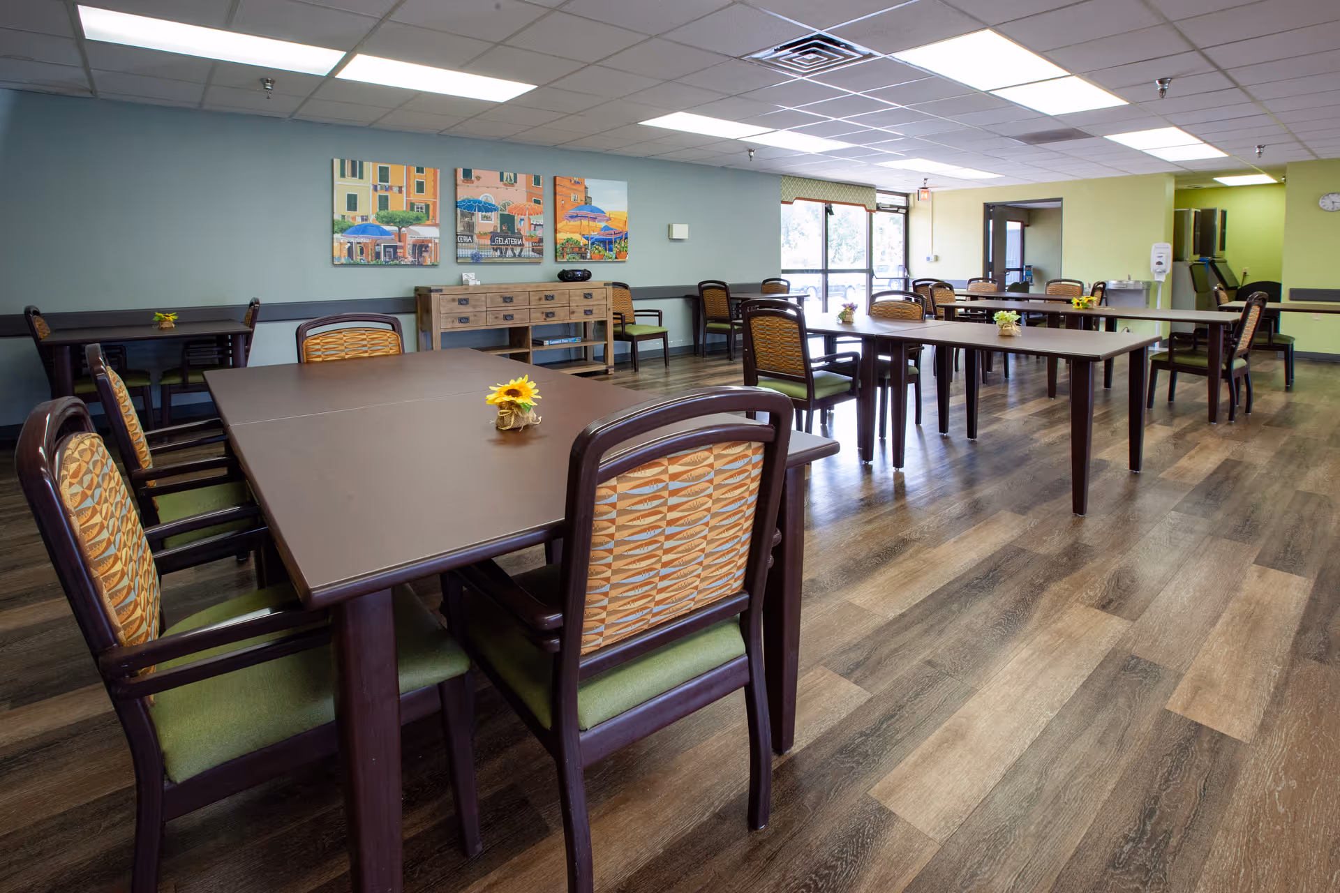 A spacious dining room with multiple dark wood tables and chairs with patterned backs and green cushions. Small flower arrangements are placed on the tables. The room has wood flooring, light green and blue walls, and a ceiling with fluorescent lighting. There are large windows letting in natural light and colorful paintings on one wall.