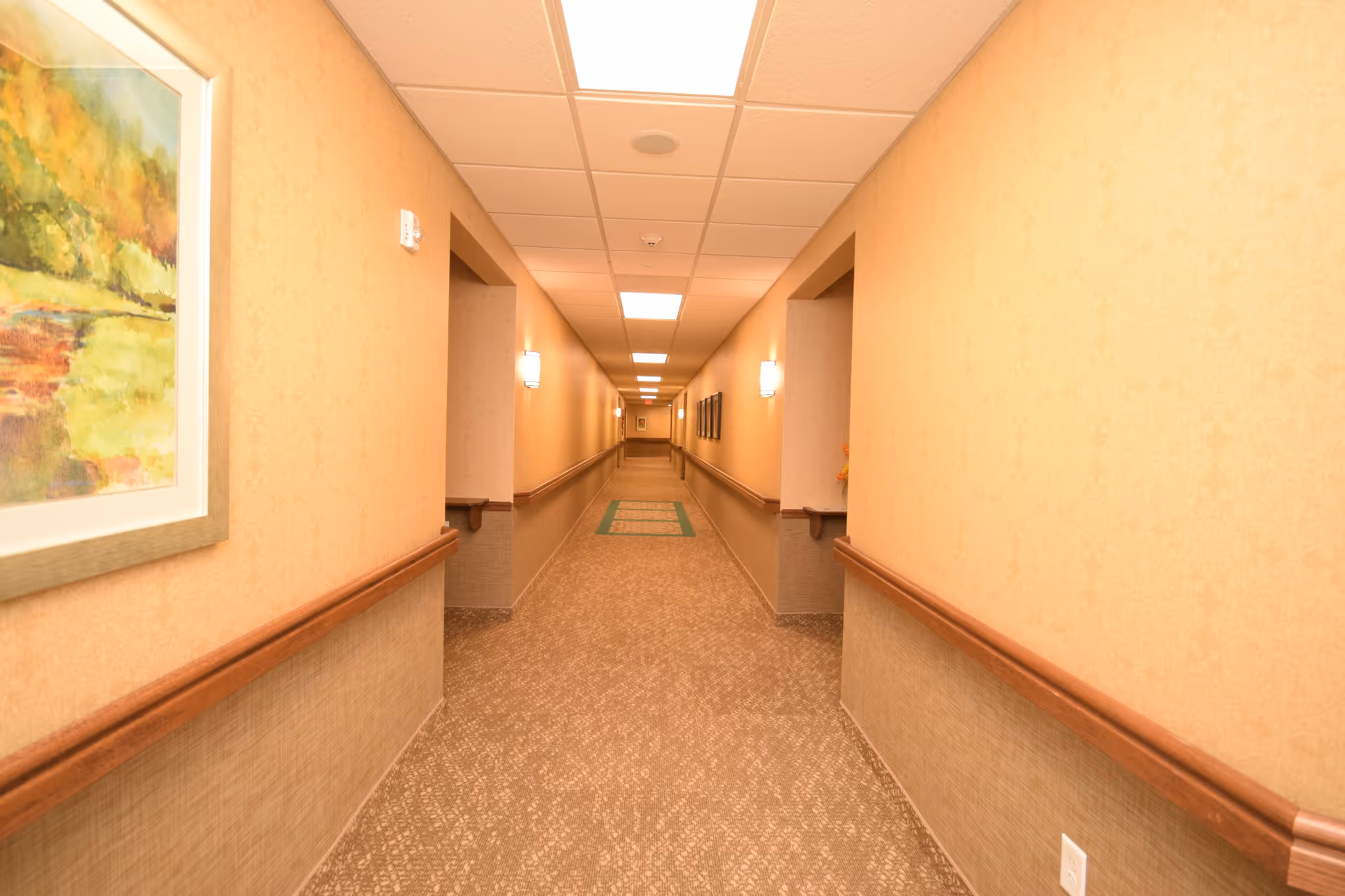 A long, well-lit hallway in a senior living facility with beige walls, carpeted floor, wooden handrails on both sides, and framed artwork on the walls.