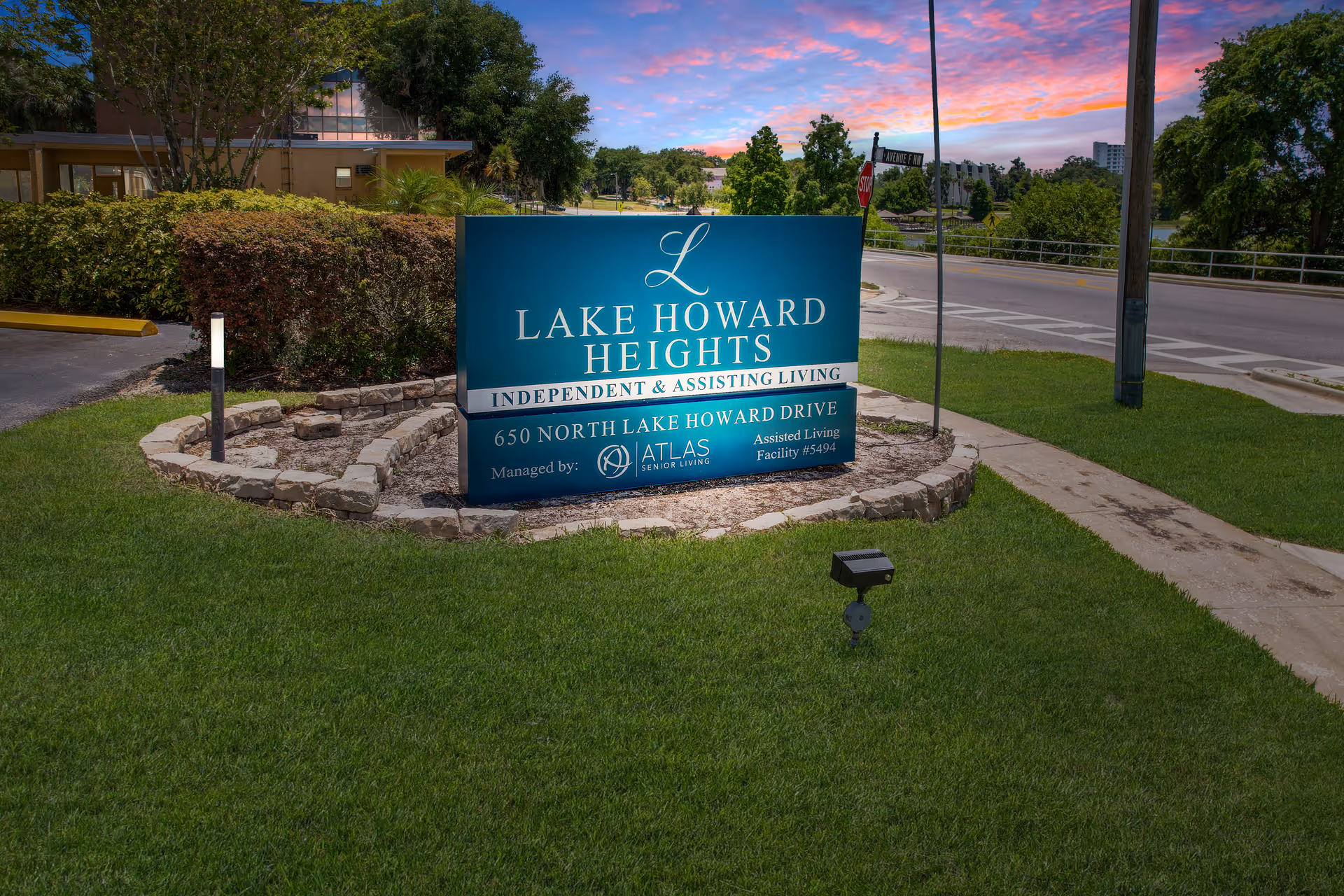 Outdoor view of a blue sign for Lake Howard Heights, an independent and assisting living facility located at 650 North Lake Howard Drive. The sign is surrounded by a small stone border and green grass, with a street and trees in the background under a colorful sunset sky.