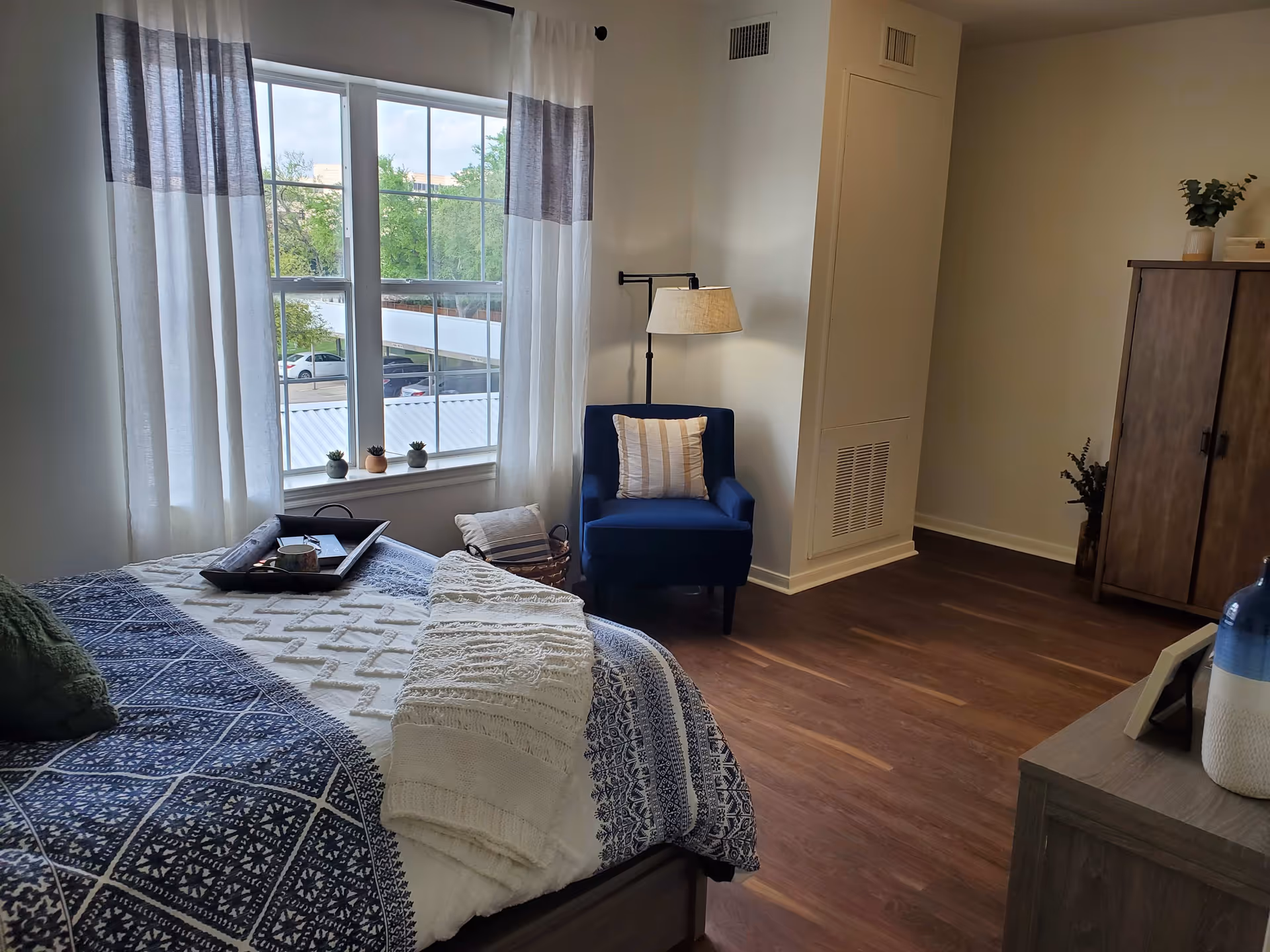 A cozy bedroom with a large window covered by white and gray curtains. The bed is made with a blue and white patterned blanket and a white knitted throw. There is a dark blue armchair with a striped pillow next to a floor lamp. A wooden cabinet and a dresser with decorative items are also visible. The room has wooden flooring and white walls.