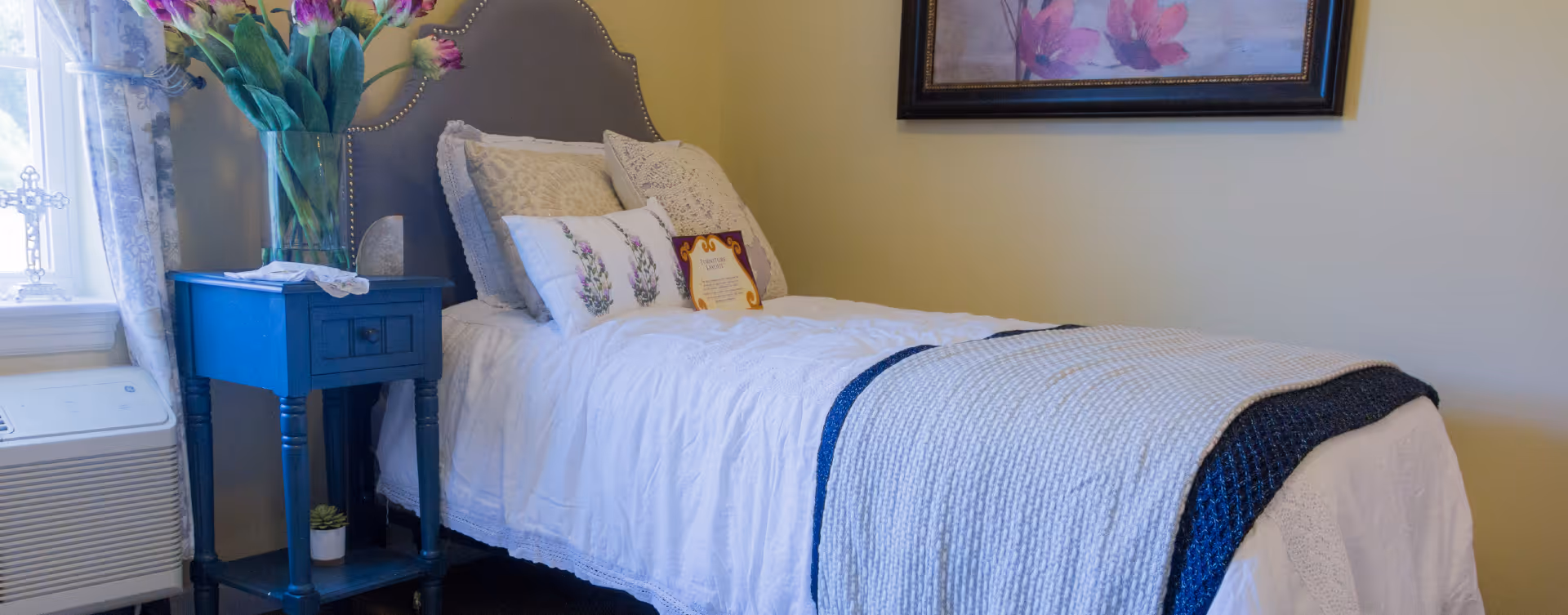 A cozy bedroom with a single bed featuring a gray upholstered headboard, white bedding, and decorative pillows. Next to the bed is a blue nightstand with a vase of purple flowers and a small potted plant. A window with patterned curtains allows natural light into the room, and a framed floral painting hangs on the wall above the bed.