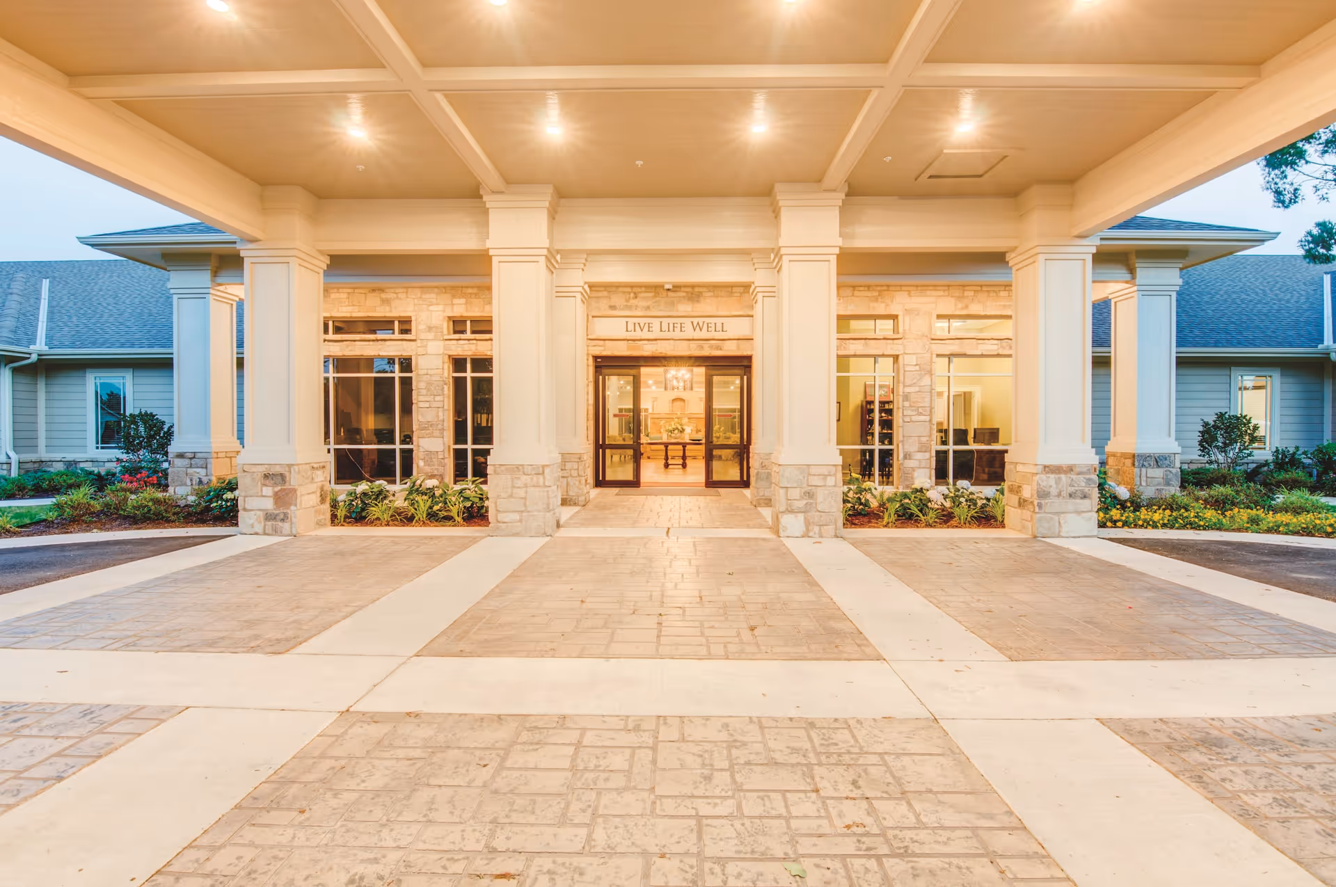 Entrance of a senior living facility with a covered driveway supported by large white columns. The building features stone and beige siding with large windows and a sign above the door that reads 'LIVE LIFE WELL'. There are landscaped plants and flowers on either side of the entrance.