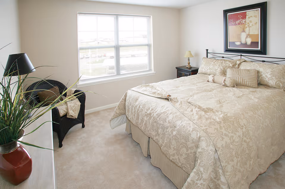 Bright, neutral-toned bedroom featuring a large bed with patterned bedding, a bedside table, and a wicker chair by a window.