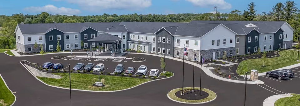 Aerial view of a two-story senior living building with a circular driveway, parking lot, flagpoles, and landscaped grounds.