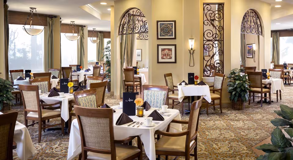 A well-lit dining room with multiple tables covered in white tablecloths, each set with folded brown napkins, cups, and silverware. The room features wooden chairs with cushioned seats and backs, decorative wall art, wrought iron wall accents, and large windows with curtains allowing natural light to fill the space. There are also small flower arrangements on each table.