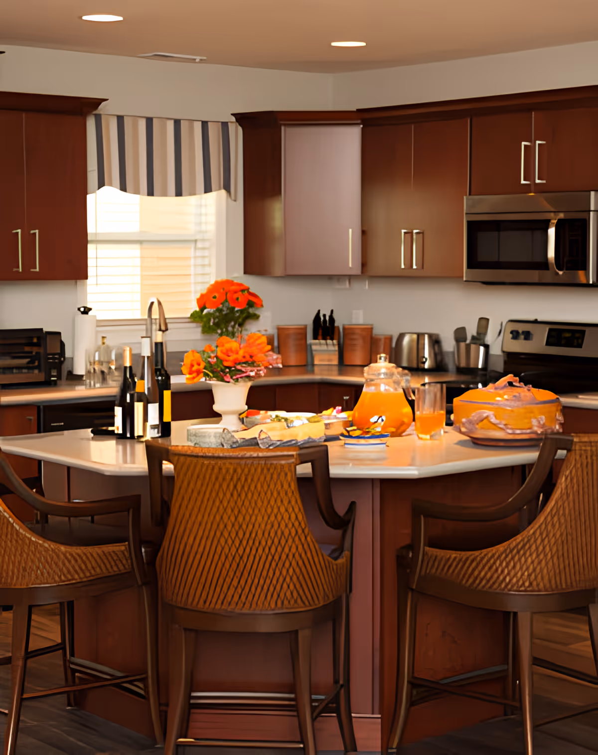 A modern kitchen with dark wood cabinets, a central island with a white countertop, three wicker bar stools, a vase of orange flowers, bottles of wine, a glass pitcher with orange juice, and various kitchen appliances including a microwave, toaster, and stove.