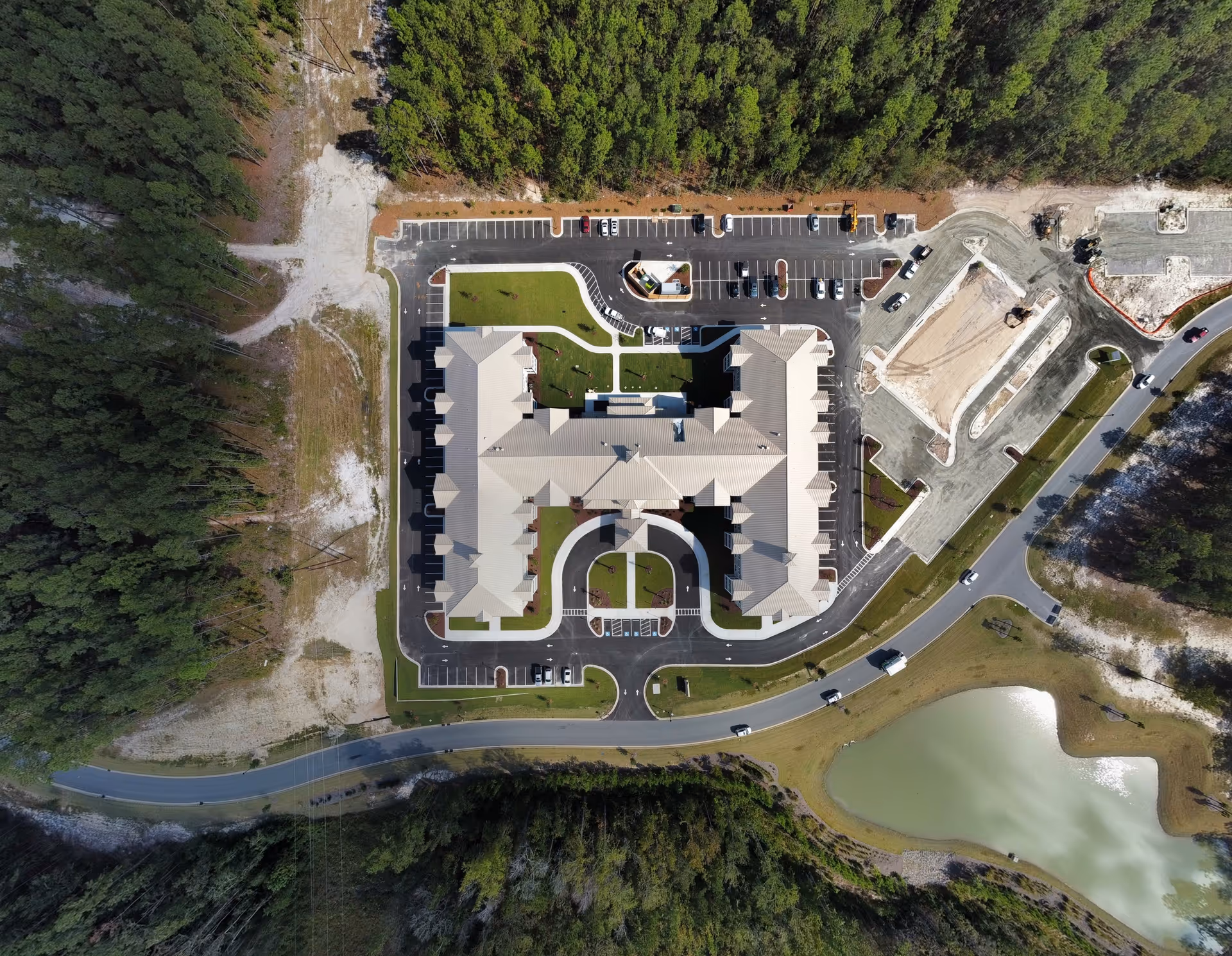 Aerial view of Arbor Landing at Compass Pointe senior living facility showing a large building with a U-shaped layout surrounded by parking lots, roads, a small pond, and dense forested areas.