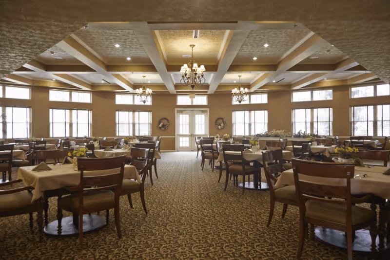 A spacious dining room with multiple tables covered in beige tablecloths and set with napkins and small floral centerpieces. The room features large windows allowing natural light, chandeliers hanging from a coffered ceiling, and a patterned carpeted floor.