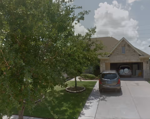 A suburban house with a two-car garage, a driveway with a parked car, and a green lawn with trees in front under a partly cloudy sky.