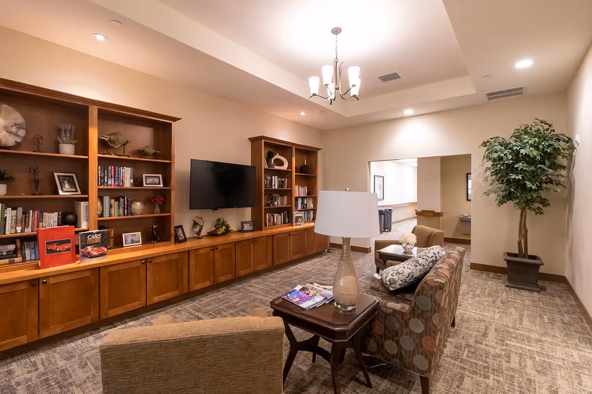 A cozy living room area in an assisted living facility featuring a wall-mounted TV, wooden bookshelves filled with books and decorative items, comfortable armchairs, a side table with a lamp, and a large potted plant in the corner. The room has soft lighting and carpeted flooring.
