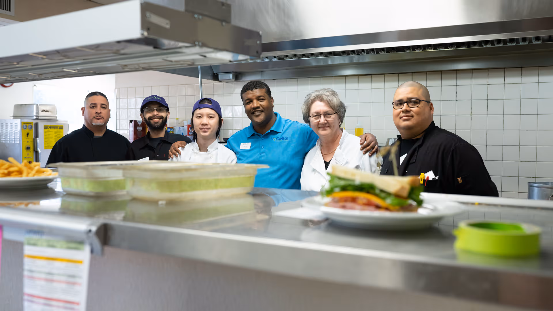 A group of kitchen staff stand behind a stainless-steel serving counter with plated food in a commercial kitchen.