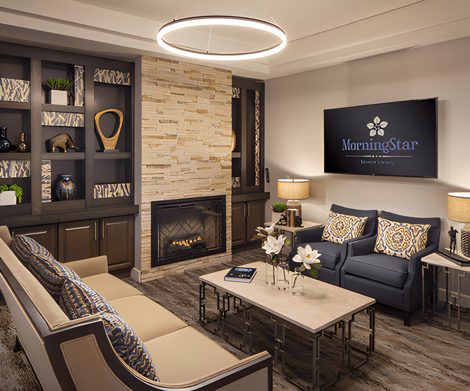 A cozy living room area in MorningStar Assisted Living & Memory Care of Rio Rancho featuring a beige sofa with patterned cushions, two dark armchairs with matching cushions, a rectangular coffee table with decorative flowers and a book, a stone fireplace with a fire burning, built-in dark wood shelves with decorative items, two side tables with lamps, and a wall-mounted TV displaying the MorningStar logo.