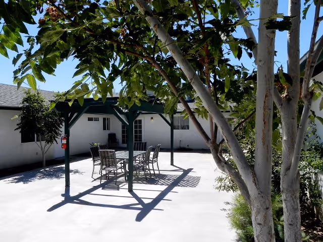 Outdoor patio area with a table and six chairs under a green pergola. The patio is surrounded by a white building with windows and doors. Trees and plants are visible around the patio providing some shade.