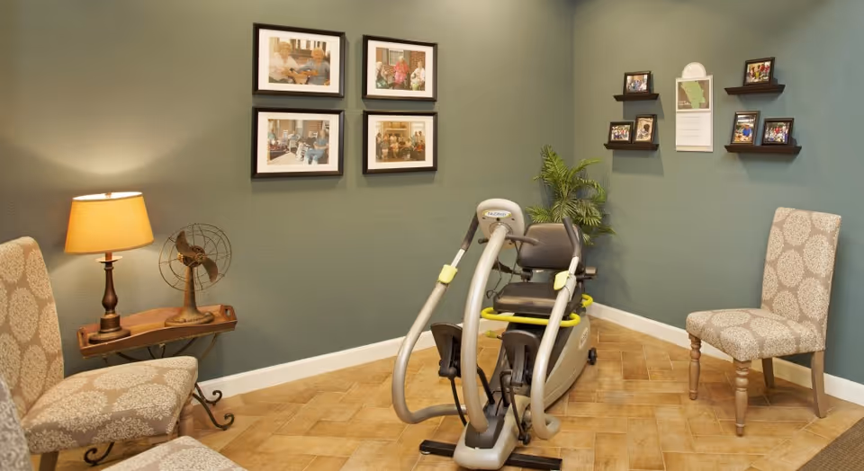 A cozy room with two patterned chairs, a small wooden side table with a lamp and vintage fan, a recumbent exercise bike, and framed photos on green walls along with small shelves displaying more pictures and a calendar.