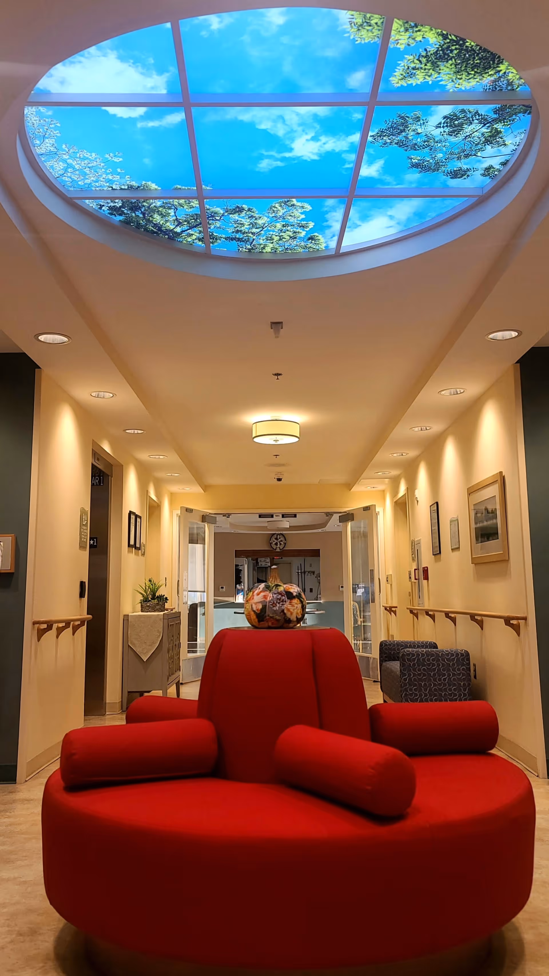 Interior hallway of AlfredHouse Symphony with a circular red cushioned bench in the center. The ceiling features a large oval skylight with an image of a blue sky and tree branches. The hallway is softly lit with recessed lights and wall sconces, and there are handrails along the walls. A small table with a plant and framed pictures are visible along the walls.