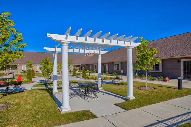 Outdoor courtyard area at Charter Senior Living of Bay City featuring a white pergola with a round metal table and chairs underneath. Surrounding the pergola are green lawns, small trees, and shrubs, with a single-story building in the background under a clear blue sky.