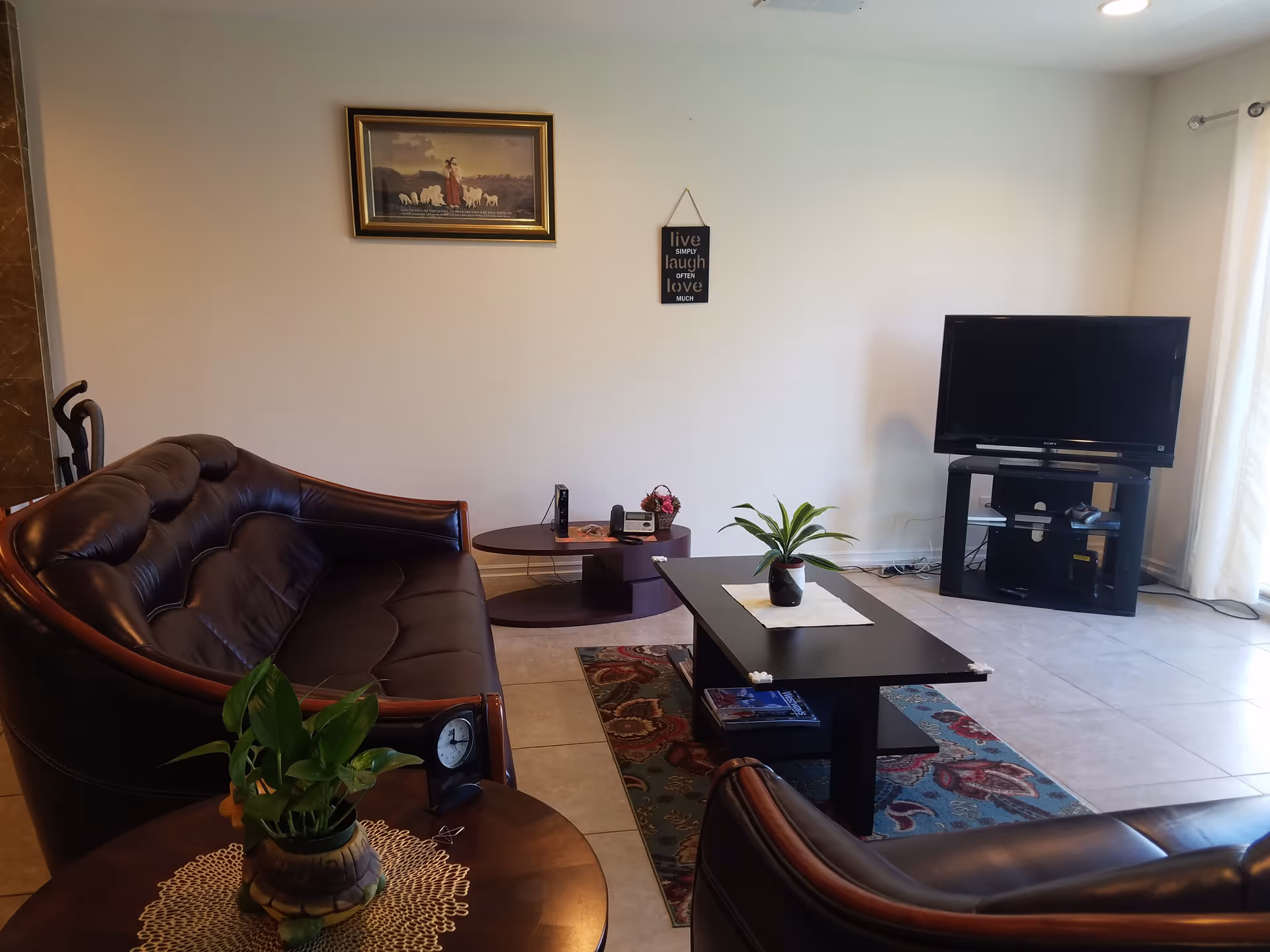 A living room with two dark brown leather sofas, a black coffee table with a small potted plant on a white cloth, a patterned rug underneath, and a TV on a black stand near a window with white curtains. There is a small round side table with a green plant and a clock, and a framed picture and a wall hanging with the words 'live simply laugh often love much' on the white wall.