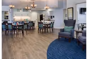 Spacious communal dining and lounge area with round tables and chairs, a kitchenette in the back, and armchairs on blue carpet.
