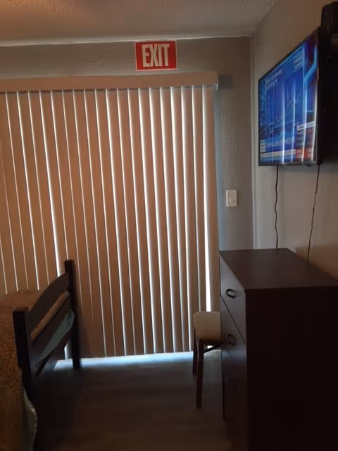 A small room with a bed on the left side, a wooden dresser with three drawers on the right, a small chair next to the dresser, vertical blinds covering a sliding glass door, and an exit sign above the door. A flat-screen TV is mounted on the wall above the dresser.