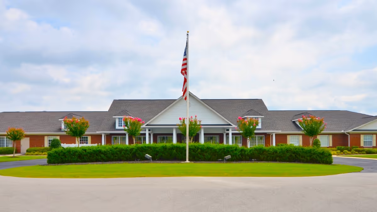 Front exterior view of The Terrace at Priceville senior living facility with a large American flag on a flagpole in the center, surrounded by neatly trimmed bushes and small trees with pink flowers, under a partly cloudy sky.