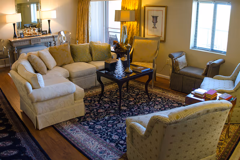 A cozy living room with a sectional sofa, several armchairs, a central coffee table on a patterned rug, and lamps by the windows.