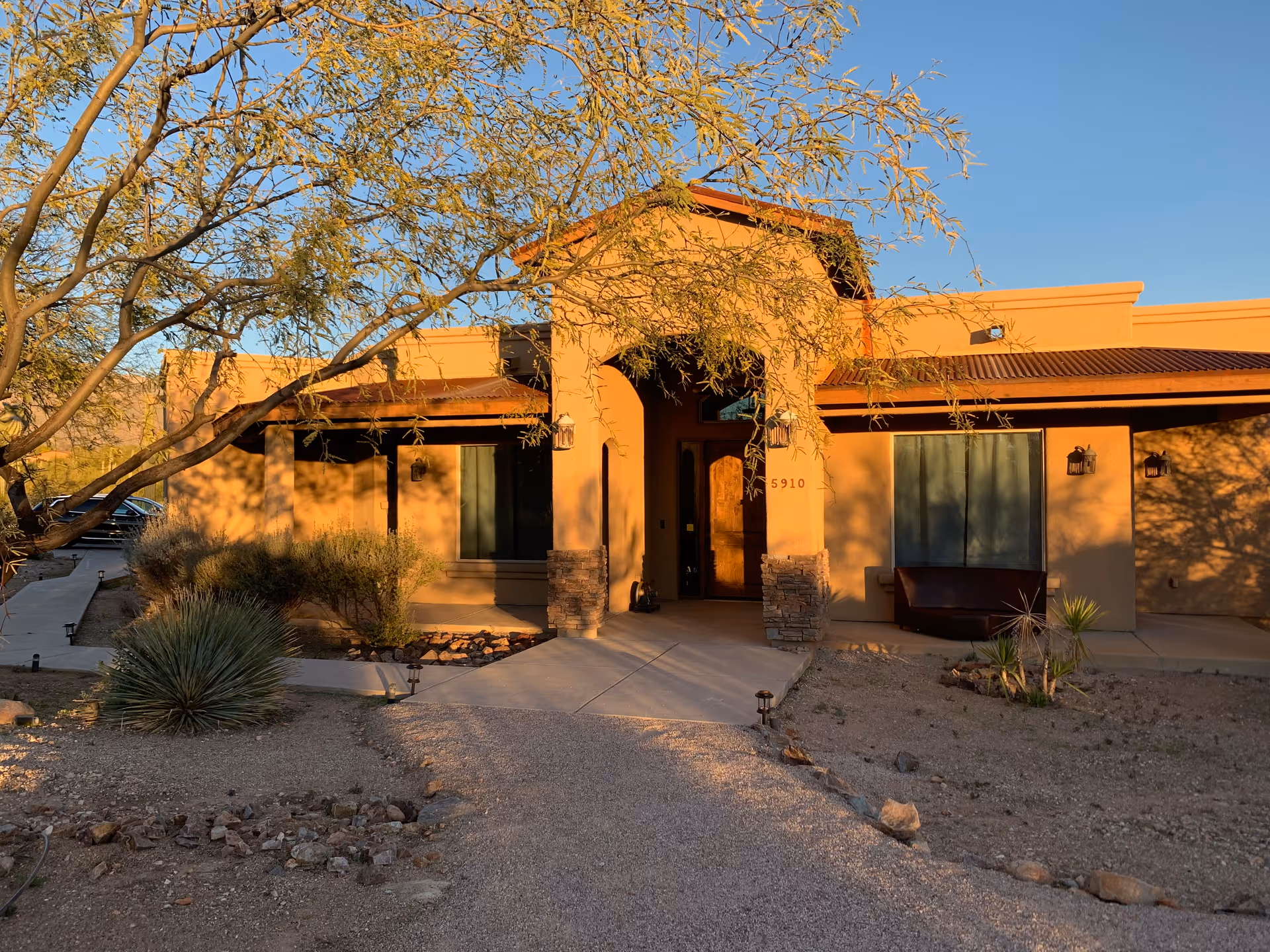 Sunlit single-story desert-style assisted living building entrance with a walkway, desert landscaping, and an overhanging tree.