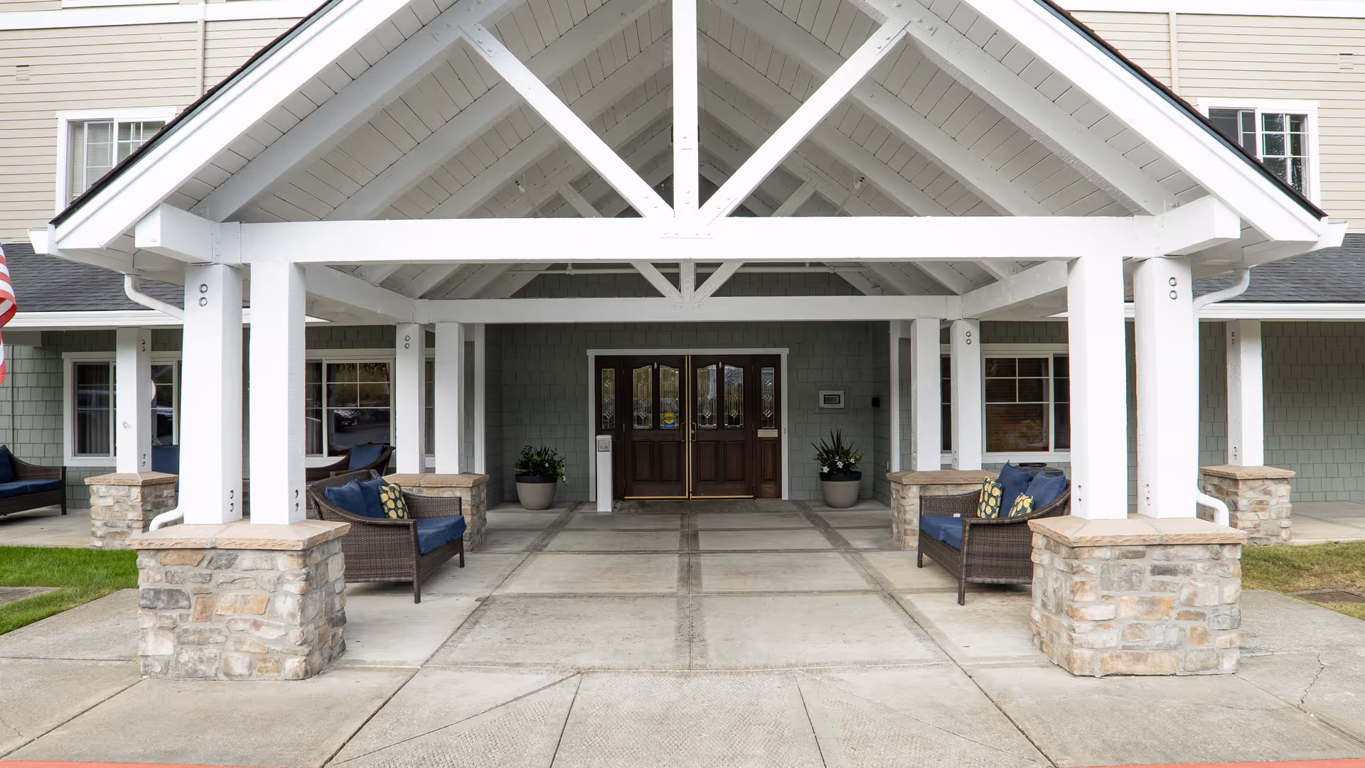 Covered entrance of a senior living facility with white wooden beams and stone pillars. There are two sets of double wooden doors in the center, flanked by potted plants. Outdoor seating with cushioned chairs is placed on either side under the covered area.