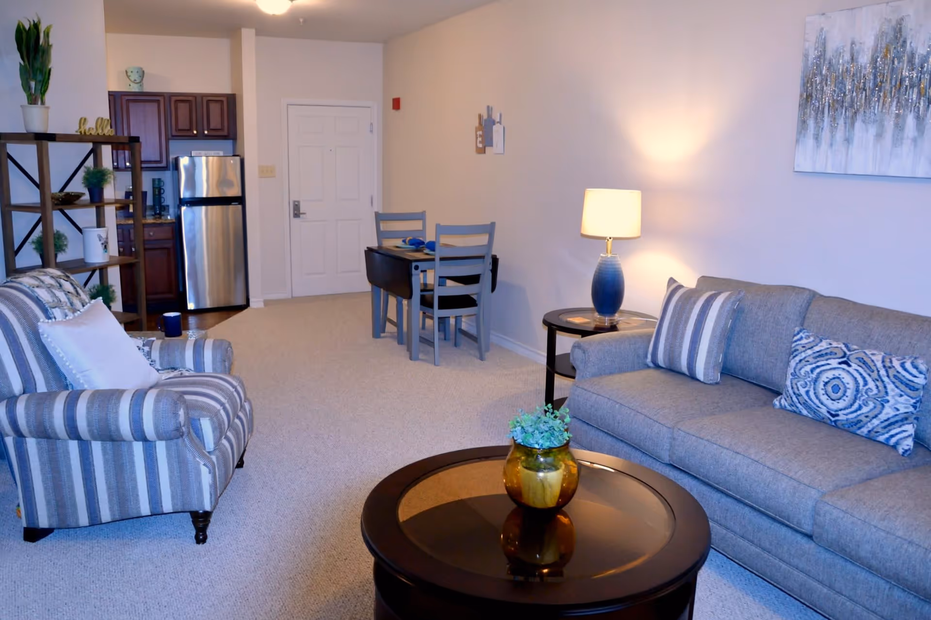 A cozy living room area with a gray sofa adorned with patterned pillows, a striped armchair with a white pillow, and a round wooden coffee table with a small plant in a yellow pot. A side table with a blue lamp is next to the sofa. In the background, there is a small dining table with two chairs and a kitchen area with dark wood cabinets and a stainless steel refrigerator. The walls are light-colored, and a decorative painting hangs above the sofa.
