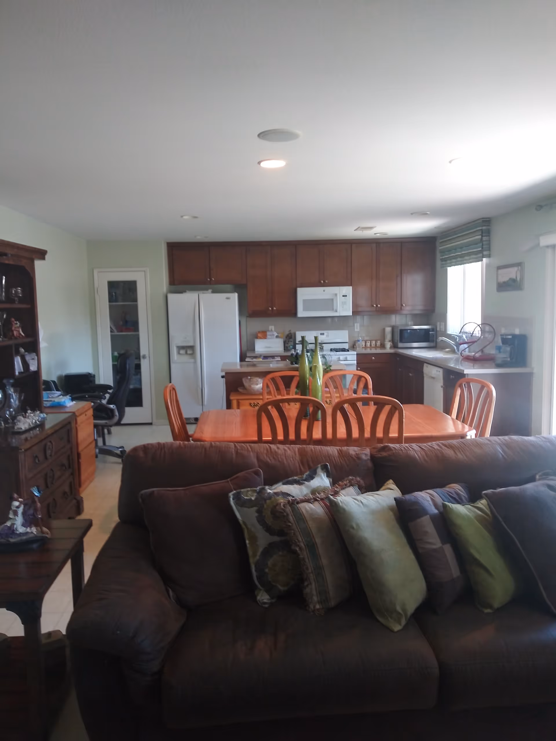 Open-plan living room with a brown sofa and decorative pillows facing a wooden dining table and a kitchen with cabinets and appliances in the background.
