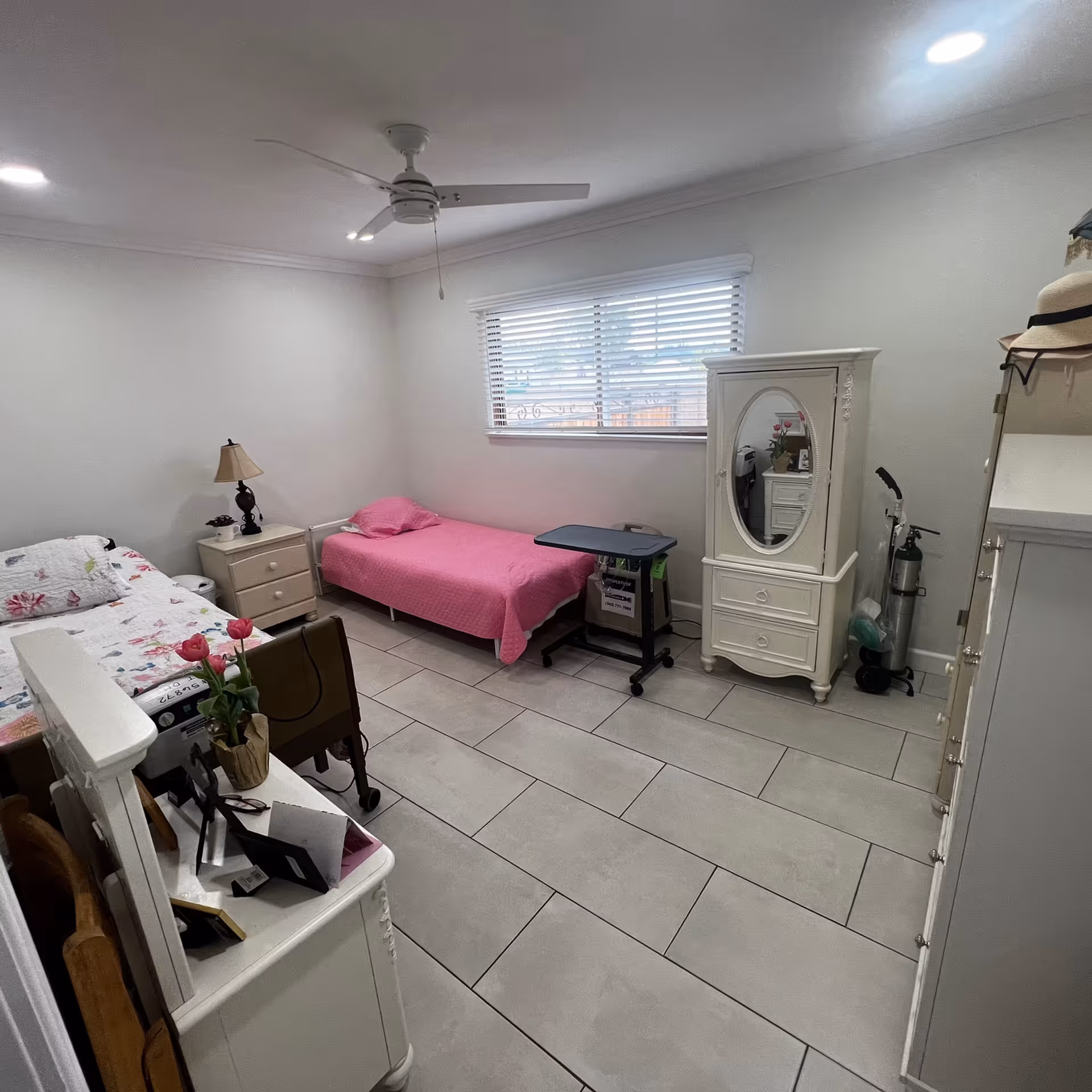 Well-lit bedroom with two single beds (one covered in pink), white furniture, a ceiling fan, tiled floor, and a window with blinds.