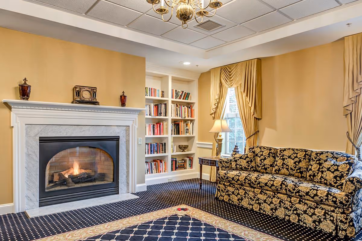 A cozy living room with a lit fireplace surrounded by a white mantel and marble trim. To the right, there is a floral patterned sofa and a side table with a lamp. Behind the sofa, there are two windows with gold drapes. A built-in bookshelf filled with books is next to the fireplace. The room has a patterned carpet and warm beige walls.