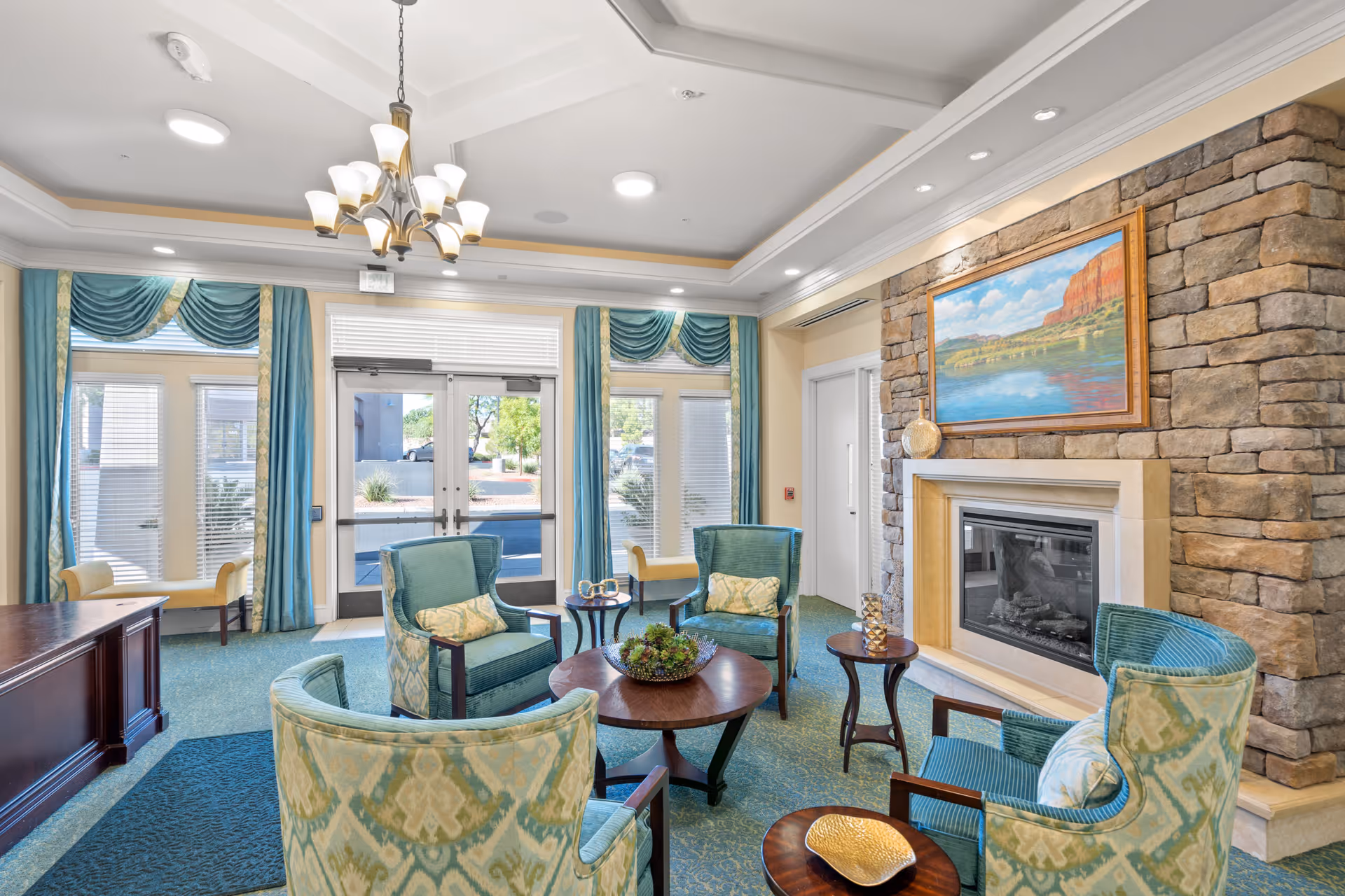 A cozy sitting area in a senior living facility with four upholstered armchairs arranged around a round wooden coffee table. The room features a stone fireplace with a landscape painting above it, large windows with teal and gold curtains, and a chandelier hanging from the ceiling. There are additional small tables with decorative items and a wooden desk visible on the left side.