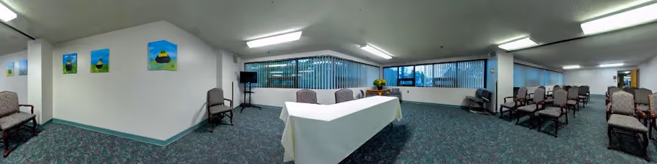 A large, carpeted common room with multiple rows of upholstered chairs arranged facing a table covered with a white tablecloth. The room has white walls adorned with colorful paintings and large windows with vertical blinds letting in natural light. There is a microphone stand near the window and a small table with a flower arrangement.