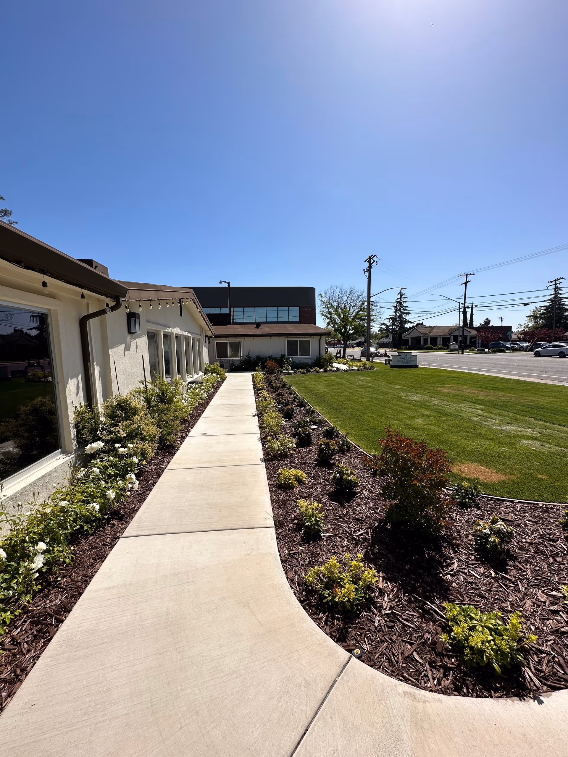 Concrete walkway beside a single-story building with landscaped beds and a grassy lawn under a clear blue sky.